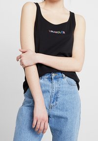 Svart linne med regnbågsfärgad "WRANGLER"-logotyp, med rundad halsringning. Matchas med höga ljusblå jeans.