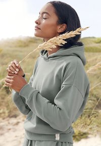 Sudadera verde claro hecha de una tela suave y texturizada con un corte relajado. Cuenta con una capucha con cordón y puños acanalados. Fondo neutro.