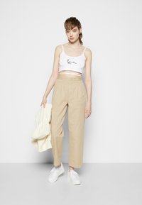 Biały krótki top na ramiączkach z czarnym napisem, beżowe spodnie z wysokim stanem i białe sneakersy. Model trzyma kremowy sweter. Neutralne tło.