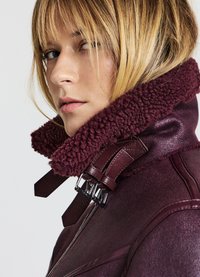 Veste en cuir bordeaux avec un col pelucheux et texturé. Dotée de quincaillerie métallique, d'une fermeture éclair et d'une coupe structurée.