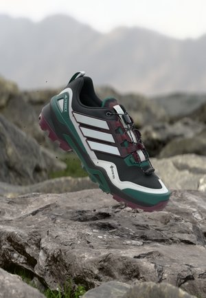 Trailrunningschoen in zwart, wit, groen en kastanjebruin, zwevend boven rotsachtig terrein met wazige bergen op de achtergrond.