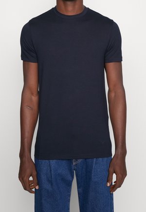 Homme portant un t-shirt bleu marine uni à manches courtes et un jean bleu, debout avec les bras détendus le long du corps sur un fond clair.