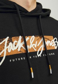 Sudadera negra con un logo frontal en blanco y naranja, con cordones que tienen acentos en naranja, negro y blanco, y un patrón texturizado.