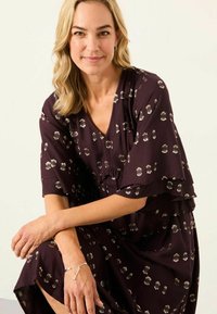 Lila Kleid mit V-Ausschnitt, rüschenförmigen Ärmeln und metallisch gemusterten Akzenten. Weicher Stoff mit einem lockeren, fließenden Design. Armband sichtbar am Handgelenk.