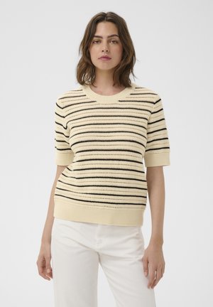 Vrouw draagt een crème kleurige korte mouwen gebreide trui met zwarte horizontale strepen en witte broek, staand tegen een lichtgrijze achtergrond.