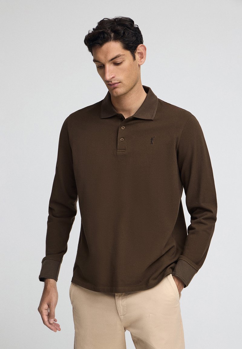Brun långärmad polo som har en krage, tre knappar fram och en liten broderad logotyp på vänster bröst. Slät tygstruktur.