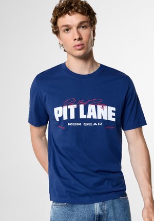 Млад мъж с къдрави коси, облечен в синя тениска с текст "Pit Lane" и "RBR Gear", комбинирана със светлосини дънки, стои на едноцветен фон.