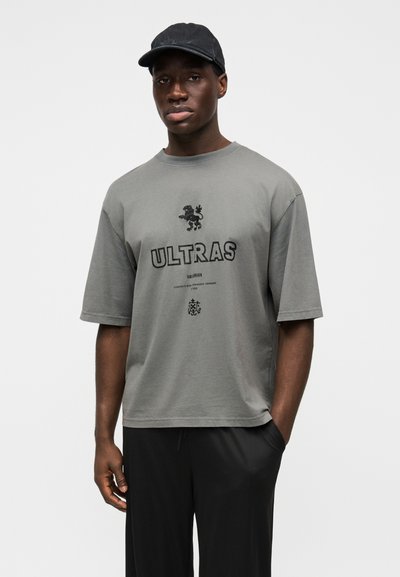 Sivá nadmerná tričko s čiernym textom "ULTRAS", obrázkom leva a tlačenými detailmi. Kombinované s čiernymi nohavicami a čiernou čiapkou.