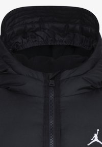 Chaqueta negra con capucha y un acabado mate liso, que presenta un cierre central con cremallera y un logo blanco en el pecho. Cuello acolchado para mayor calidez.