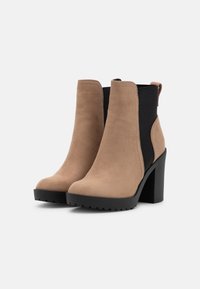 Botines beige de ante con un acabado liso, paneles laterales elásticos negros y un tacón grueso negro. Suela texturizada para mayor agarre.