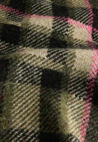 Nahaufnahme eines strukturierten Wollstoffs mit einem olivgrünen, cremefarbenen, schwarzen und rosa Karomuster, das diagonale Linien und Kästchen bildet.