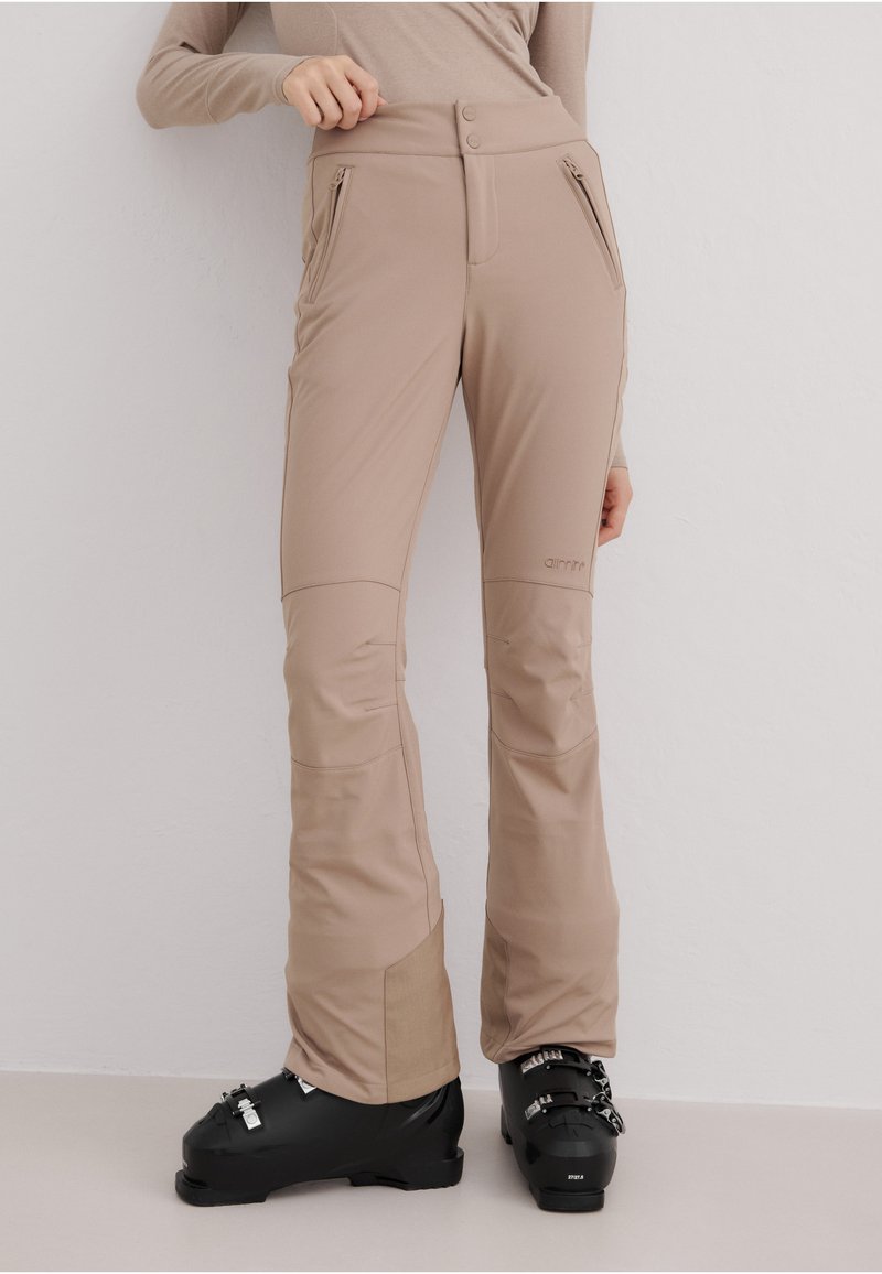Beige skibukser med høy midje, to glidelåslommer og forsterkede paneler ved knærne. Kombinert med svarte skistøvler.
