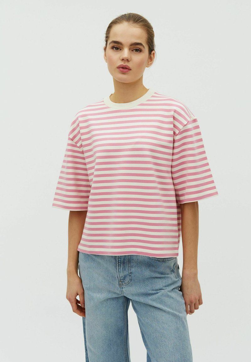 Jonge vrouw die een roze-wit gestreept T-shirt met korte mouwen en lichtblauwe spijkerbroek draagt, staand tegen een effen achtergrond.