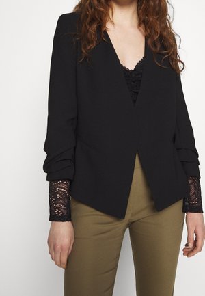 Femme portant un blazer noir sur un top en dentelle noire, associé à un pantalon vert olive, visible des épaules aux hanches sur un fond uni.