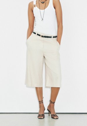 Frau trägt ärmelloses weißes Oberteil, beige Culottes mit weitem Bein und schwarzem Gürtel, schwarze Riemchen-Hochsandalen und geschichtete Halsketten, Hände in den Taschen.