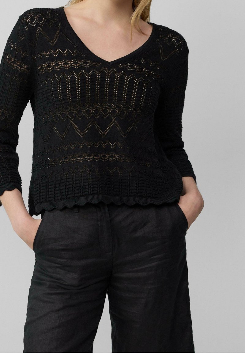 Femme portant un haut en dentelle noire à motif avec des manches trois-quarts et un pantalon taille haute noir, mains dans les poches, devant un fond gris.