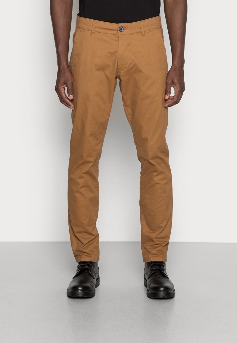 Chinos de algodón marrón con un ajuste ajustado, bolsillos frontales y un cierre de botón. Combinados con zapatos negros de cordones para un look completo.