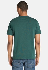 Muž v tmavě zeleném tričku s krátkým rukávem a modrých džínech, zobrazený zezadu na jednobarevném bílém pozadí.