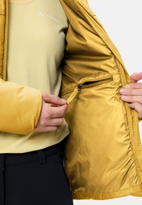 Giacca isolante gialla con una texture liscia e lucida. L'interno presenta una tasca con zip e un'etichetta in tessuto. Indossata sopra una maglia verde chiaro.