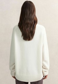 Biely oversized sveter s rebrovanými manžetami a lemom, s jemnou textúrou a voľným strihom. Pohľad zozadu, vlasy padajúce dolu.