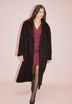 Temno volneno plašč z širokimi reverji in dvojno zaklopnimi gumbi. Nosi se preko burgundijske obleke z gumboma spredaj. Gladka textura.