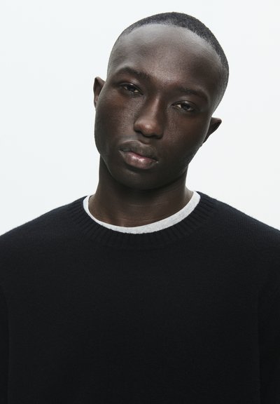 Juodas megztinis su pienišku apykaklės kraštu, dėvimas virš balto marškinio. Lygus paviršius, prigludusi forma ir neutrali aplinka.