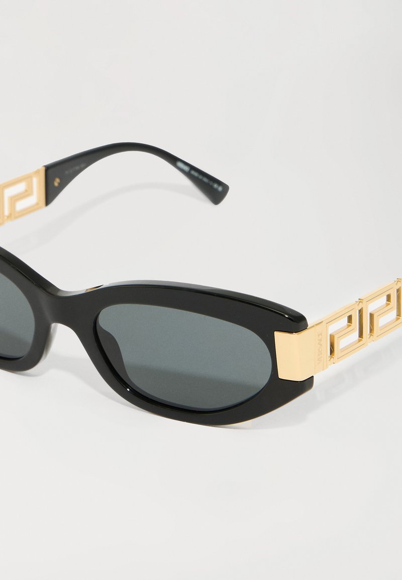 Zwarte zonnebril met een slank, ovaal frame, voorzien van donkergrijze lenzen en gouden geometrische accenten op de pootjes. Duurzaam materiaal.