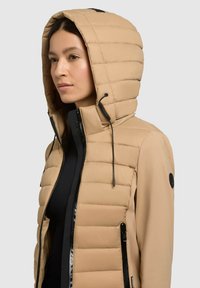 Beige polstret jakke med en glat, kontrasterende sort lynlås og detaljer. Har en quiltet hætte og en tætsiddende silhuet med sidelommer.