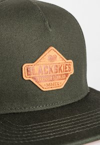 Grön keps med en texturerad, brun läderpatch som har ett emblem och orden "BLACKSKIES PREMIUM SUPPLIES" präglade. Platt skärm-design.