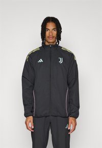 Veste d'athlétisme noire avec un col haut, présentant des accents jaunes et roses, une fermeture éclair et un logo sur la poitrine. Le matériau semble léger.