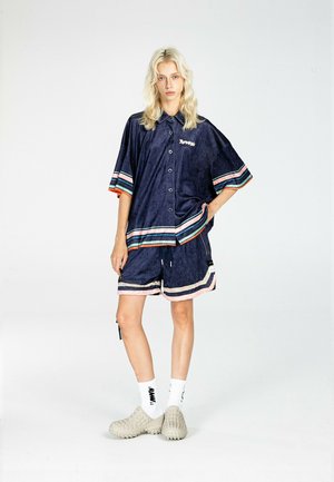 Joven mujer con cabello rubio está de pie vistiendo una camisa azul marino de botones de gran tamaño y pantalones cortos a juego con rayas multicolor, calcetines blancos y zapatillas beige.