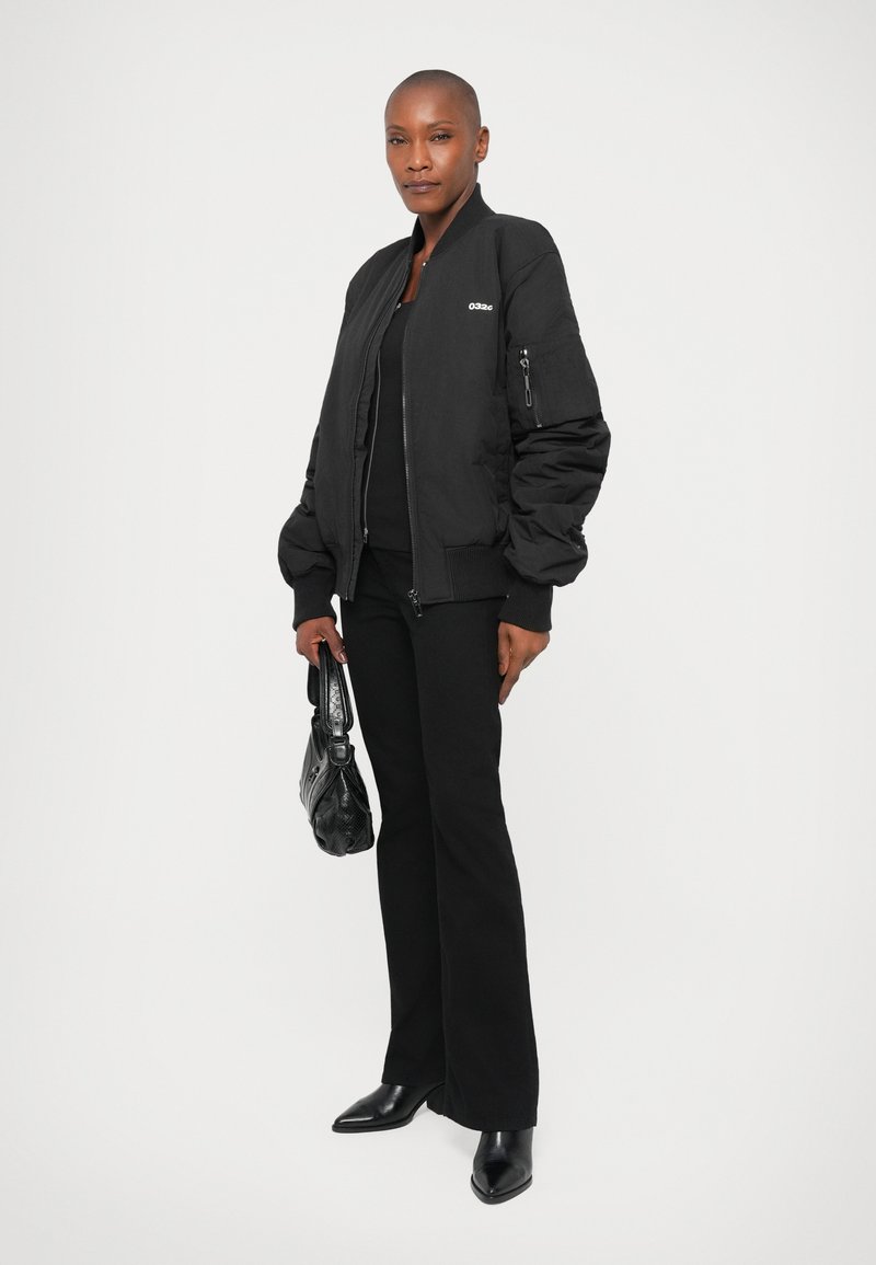 Person wearing a black bomber jacket, black pants, and black heeled boots, holding a black handbag against a plain white background.