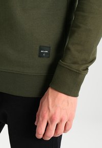 Vihreä pitkähihainen paita, jossa on ribbikuositetut hihansuut ja helma. Alareunassa on ommeltu pieni musta merkki, jossa lukee "ONLY & SONS." Lähikuva kädestä.