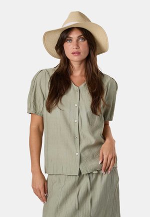 Femme aux longs cheveux bruns portant un chapeau beige à large bord, une chemise boutonnée à manches courtes vert clair et une jupe assortie, debout sur fond blanc.