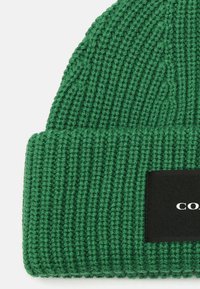 Berretto di lana verde lavorato a maglia con texture a coste e un'etichetta rettangolare nera con testo bianco sul davanti.