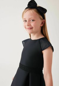 Schwarzes Kleid mit kurzen Ärmelchen, strukturierter Halslinie und einem gerafften Rock. Es hat einen schwarzen markierten Taillengürtel und ein großes schwarzes Schleifenhaarband.
