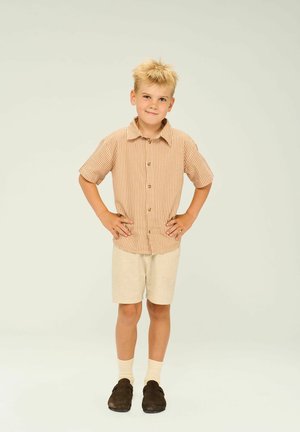Camisa de manga corta a rayas en naranja y blanco, pantalones cortos beige, calcetines beige, zapatos oscuros, de pie con las manos en la cadera frente a un fondo neutro.