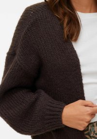 Ciemnobrązowy sweter na drutach z teksturowanym wzorem, opuszczonymi ramionami i ściągaczami przy rękawach. Materiał wydaje się miękki i ciepły.