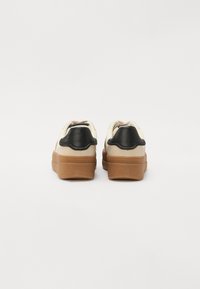 Sneakers z kremową cholewką, czarnym skórzanym akcentem na pięcie i brązową gumową podeszwą. Okrągły czubek z detalami sznurowania na górze.