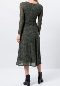 Frau in einem dunkelgrünen, langärmligen Midikleid mit gesprenkeltem Muster und schwarzen, hohen Stiefeletten, die mit dem Rücken zur Kamera steht.