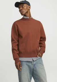 Suéter de punto marrón con cuello alto, puños acanalados y dobladillo, superpuesto a una camisa a rayas blanca y negra, combinado con jeans de color azul claro.