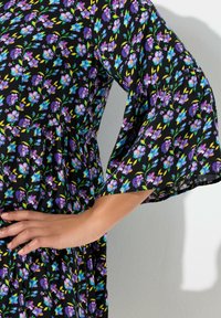 Blomsterkjole med sort baggrund med lilla og blå blomster, lange klokkeærmer og en indsnævret talje. Glat stoftekstur.