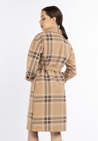 Donna con capelli scuri legati all'indietro, che indossa un cappotto beige a scacchi con cintura, in posizione laterale su uno sfondo bianco.