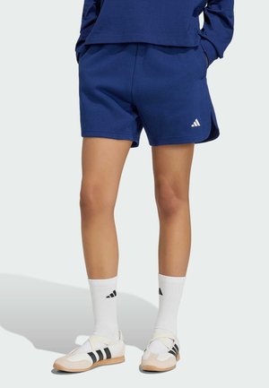 Persona che indossa pantaloncini sportivi blu navy, calzini bianchi con logo nero e scarpe da ginnastica beige con strisce nere, in piedi con le mani in tasca.