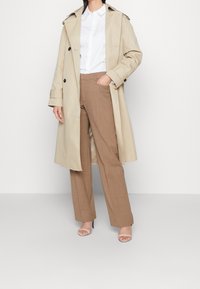 Trench-coat beige, chemise blanche à boutons, et pantalon marron ajusté, associés à des sandales à talons transparents, sur un fond uni.