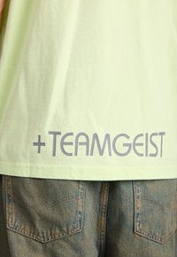 Světle zelené bavlněné tričko s hladkou texturou, na zadní straně s odvážným šedým nápisem "+TEAMGEIST". Spárováno s džínovými kalhotami s texturovanými kapsami.