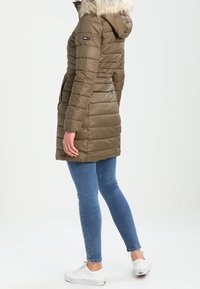 Abrigo puffer marrón con capucha forrada de pelo, textura acanalada y costuras verticales. Lleva puesto sobre unos vaqueros azules y zapatillas blancas.