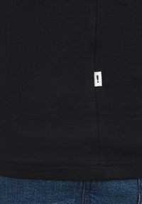 T-shirt nera in cotone con etichetta sulla cucitura laterale che presenta uno sfondo bianco e un punto esclamativo nero. Jeans in denim visibili nella parte inferiore.