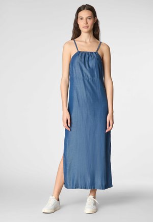 Modella che indossa un abito maxi in denimo senza maniche con scollatura arricciata e spacco laterale, abbinato a sneakers bianche, su uno sfondo neutro.