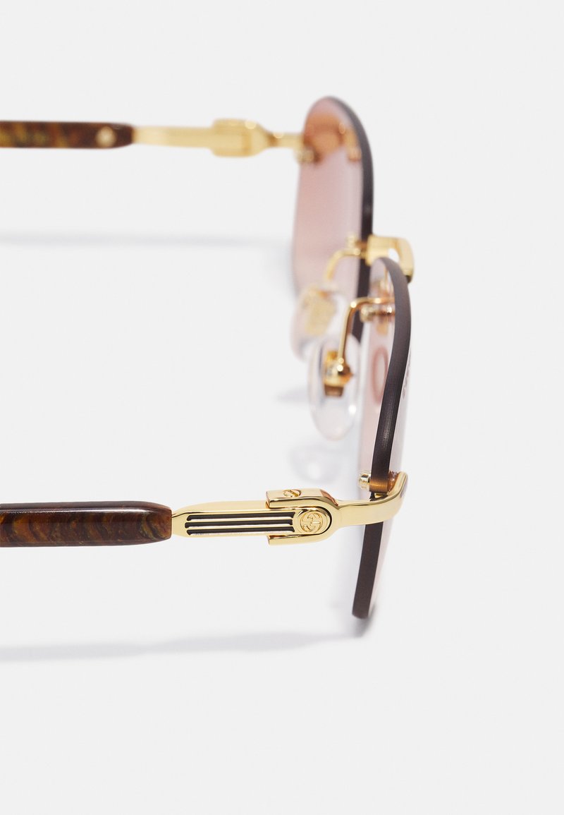 Lunettes de soleil marron et dorées avec des verres ronds, une monture en brown à motifs et des accents dorés sur la monture et la charnière. Plaquettes de nez transparentes incluses.